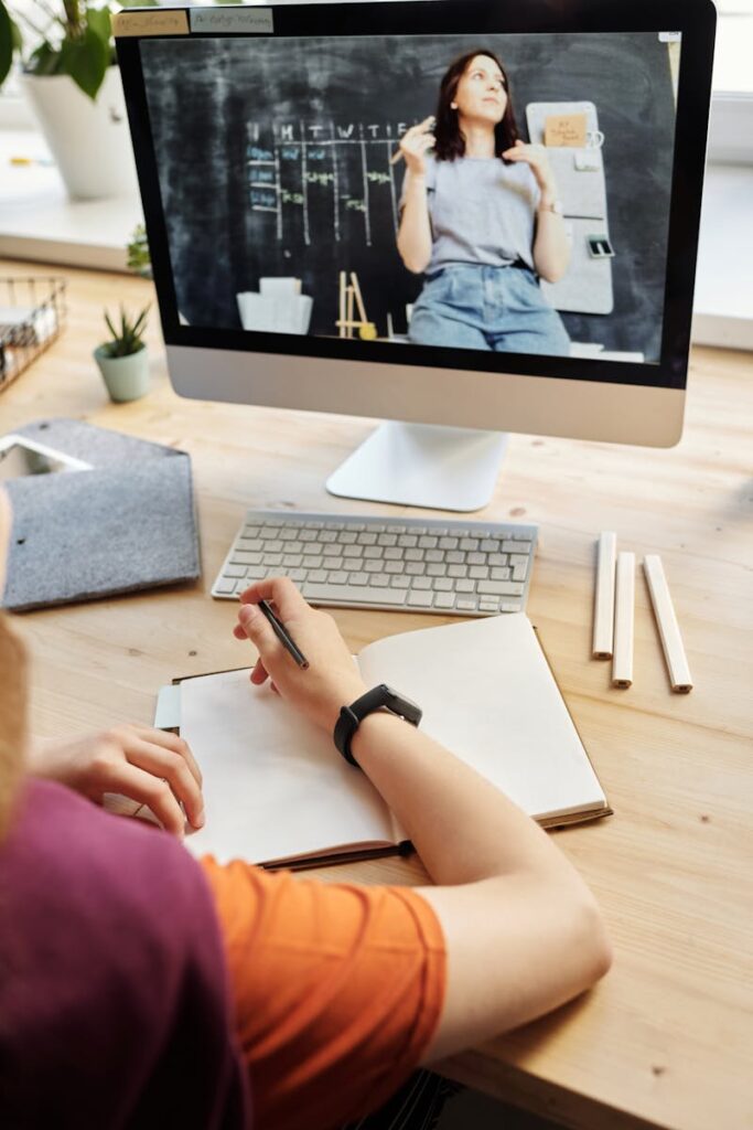 Student attending virtual class on a computer, taking notes at home.