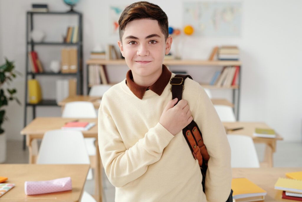 Smiling teenage boy with a backpack in a bright classroom setting.